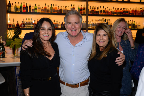 Three people smiling and posing together in a bar with illuminated shelves of liquor bottles behind them; two women stand on either side of a man with his arms around their shoulders. The lively After Hours scene is done right, with others mingling in the background. Lifestyle