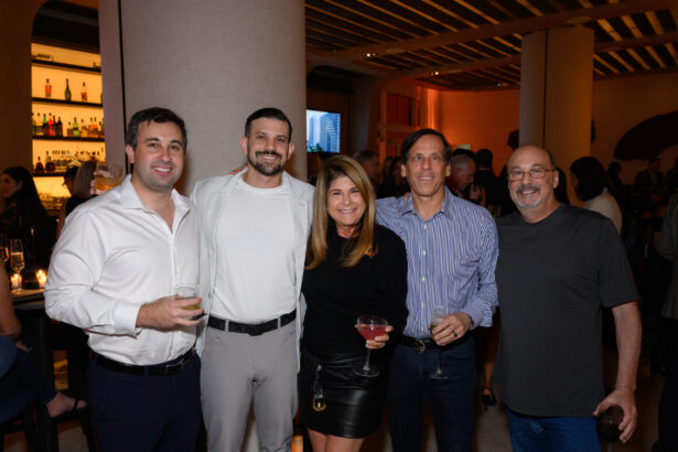 Five people stand together indoors at an After Hours social event, smiling at the camera. They are holding drinks, and the background shows a bar area with warm lighting and other guests. Lifestyle