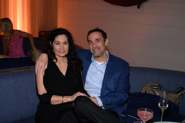 A man in a blue blazer and a woman in black sit closely together on a blue couch, smiling at the camera during an After Hours gathering. A pink cocktail and wine glass rest on the table, while another woman sits in the background. Lifestyle