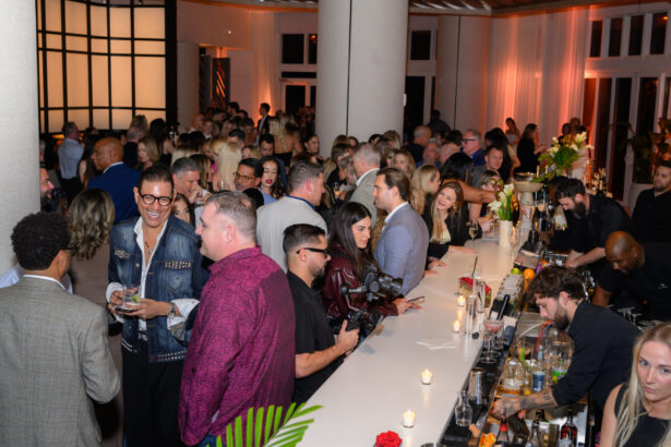 A large crowd of people socialize and mingle around a well-lit bar at an After Hours indoor event. Bartenders serve drinks as guests chat, laugh, and enjoy themselves in a stylish, modern setting. Lifestyle