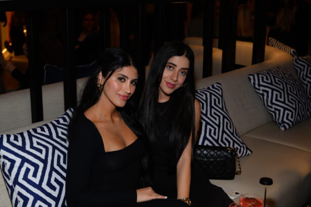 Two women with long dark hair sit closely together on a couch with blue and white geometric cushions, both in black outfits and smiling at the camera, capturing a chic After Hours moment in a dimly lit lounge or restaurant. Lifestyle