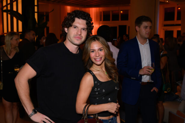 A man in a black t-shirt and a woman in a black sleeveless top pose and smile at an After Hours indoor event, with other people socializing in the background. Lifestyle
