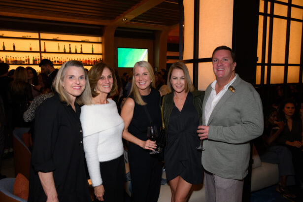 Five adults pose together at an indoor after hours social event, smiling at the camera. Two women hold glasses of wine. The background features a lit bar and other guests mingling in the modern, warmly lit space—a night done right. Lifestyle