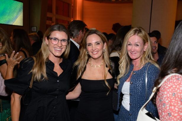 Three women smiling and posing together at an upscale After Hours social event, with warm, festive lighting and other guests mingling in the background. Lifestyle
