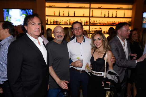 A group of four people posing together at a crowded indoor event, dressed in semi-formal attire, each holding a drink; shelves with bottles are visible in the background—an After Hours gathering done right. Lifestyle