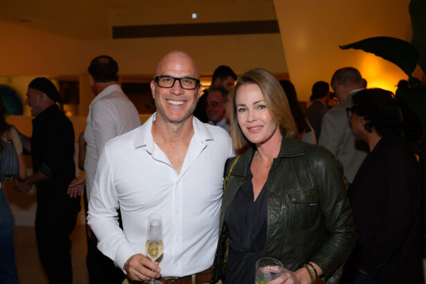 A smiling man in a white shirt and a woman in a dark jacket stand close together at an After Hours social event, each holding a drink, with other people mingling in the softly lit background. Lifestyle