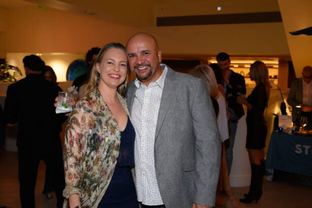A smiling woman and man pose together at an After Hours indoor event. The woman holds a drink and wears a floral jacket, while the man sports a gray blazer. Other people are mingling in the softly lit background. Lifestyle
