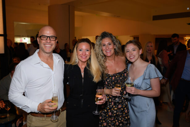 Four people stand closely together at an indoor event, smiling at the camera with glasses of wine or champagne—perfectly capturing an After Hours gathering done right. Warm lighting and other attendees fill the background. Lifestyle