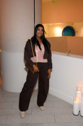 A woman with long black hair poses indoors, smiling. Dressed in a brown jacket and pants, a pale pink top, and platform shoes, she carries a brown shoulder bag—serving After Hours style done right. Candles and a drink rest on the white ledge beside her. Lifestyle