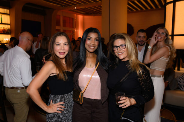 Three women stand close together and smile at the camera at an after hours social event, with other people mingling in the warmly lit, stylishly decorated space—an evening done right. Lifestyle