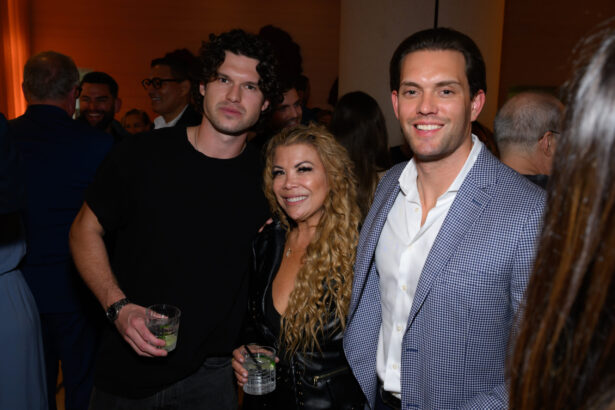 Three people pose together at an After Hours social event, smiling at the camera. The two men hold drinks while the woman stands between them. Other attendees and warm lighting appear in the background. Lifestyle