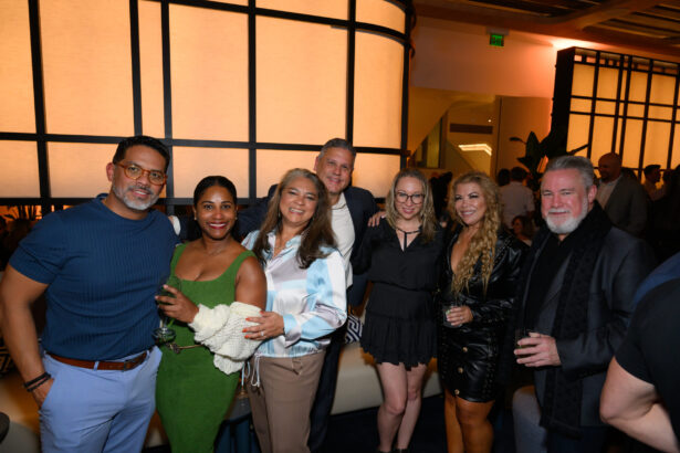 A group of seven adults, dressed in semi-formal attire, stand closely together smiling at an After Hours indoor social event; warm lighting and a modern interior are visible in the background. Lifestyle