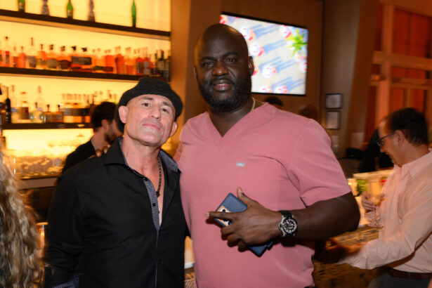 Two men pose together in a bar after hours, one wearing a black shirt and hat, the other in a pink shirt holding a phone. Shelves with bottles and a TV screen are visible in the background, with other people nearby. Lifestyle