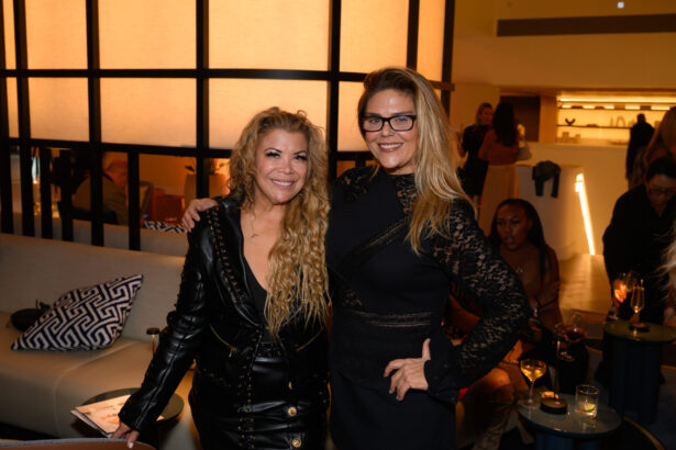 Two women smiling and posing together at a stylish indoor after hours event; one wears a black leather dress, the other a black lace dress. People sit and mingle in the background, with drinks on tables around them. Lifestyle