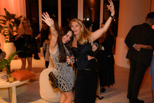 Two women pose and smile with arms raised at a lively after hours party. One wears a patterned dress, the other a black outfit. People mingle in the background, and the room is warmly lit with plants and modern decor. Lifestyle
