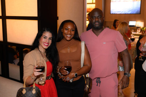 Three people pose together indoors at an After Hours social event, smiling and holding glasses of red wine. The group includes two women and one man, with other guests visible in the background. Lifestyle