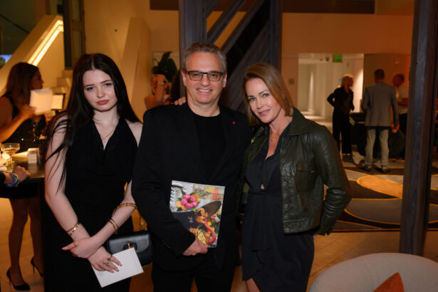 Three people pose together at an indoor event; the man in the center holds a magazine, while the two women stand on either side of him in chic evening attire. It’s an After Hours gathering done right, with guests mingling stylishly in the background. Lifestyle