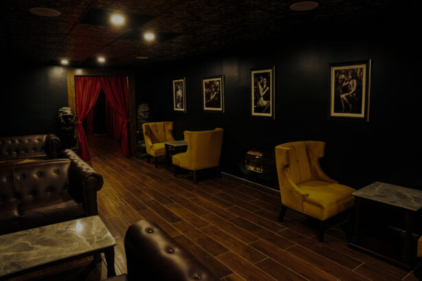 Behind the Red Door, a dimly lit lounge features dark walls, brown leather sofas, yellow armchairs, small tables, framed black-and-white photos on the wall, a wooden floor, and striking red curtains in the background. Lifestyle