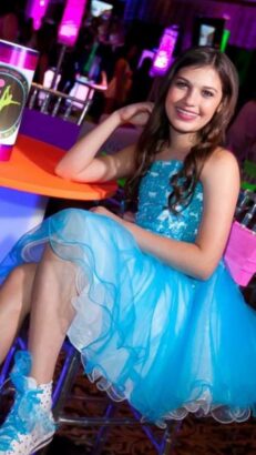 A smiling girl with long brown hair sits at a table in Fort Lauderdale, wearing a blue and white tulle dress and white sneakers. The colorful, lively background suggests she’s enjoying a joyful Inner Circle celebration or party. Lifestyle