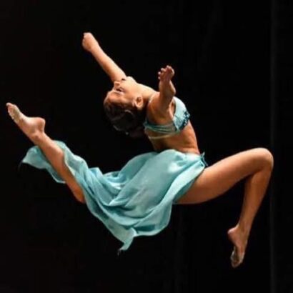 A female dancer in a flowing blue costume leaps gracefully in mid-air against a dark background, with arms and legs extended and her face turned upward, capturing the vibrant spirit of Fort Lauderdale's talented Inner Circle performers. Lifestyle