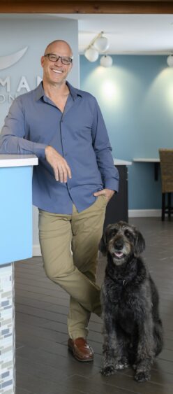 A smiling man in glasses, a blue shirt, and khaki pants leans on a blue counter in a modern health care office, with his large black and gray dog sitting beside him—a scene reflecting compassionate leaders at work. Lifestyle