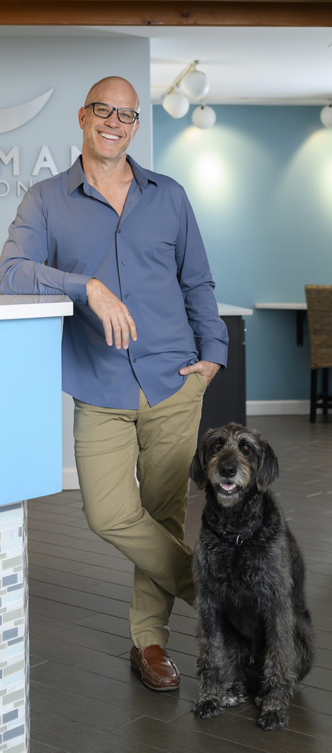 A smiling man in glasses, a blue shirt, and khaki pants leans on a blue counter in a modern health care office, with his large black and gray dog sitting beside him—a scene reflecting compassionate leaders at work. Lifestyle
