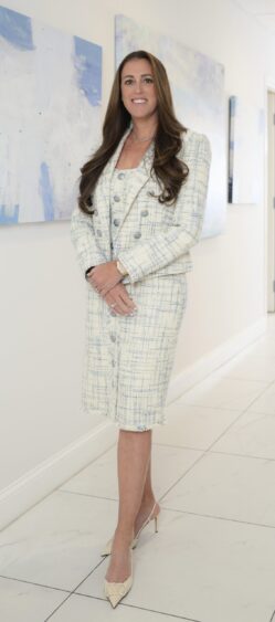 A woman with long brown hair stands indoors, smiling confidently. She wears a light-colored, textured skirt suit with buttons and nude heels. Abstract artwork hangs on the white walls behind this rising leader in health care. Lifestyle