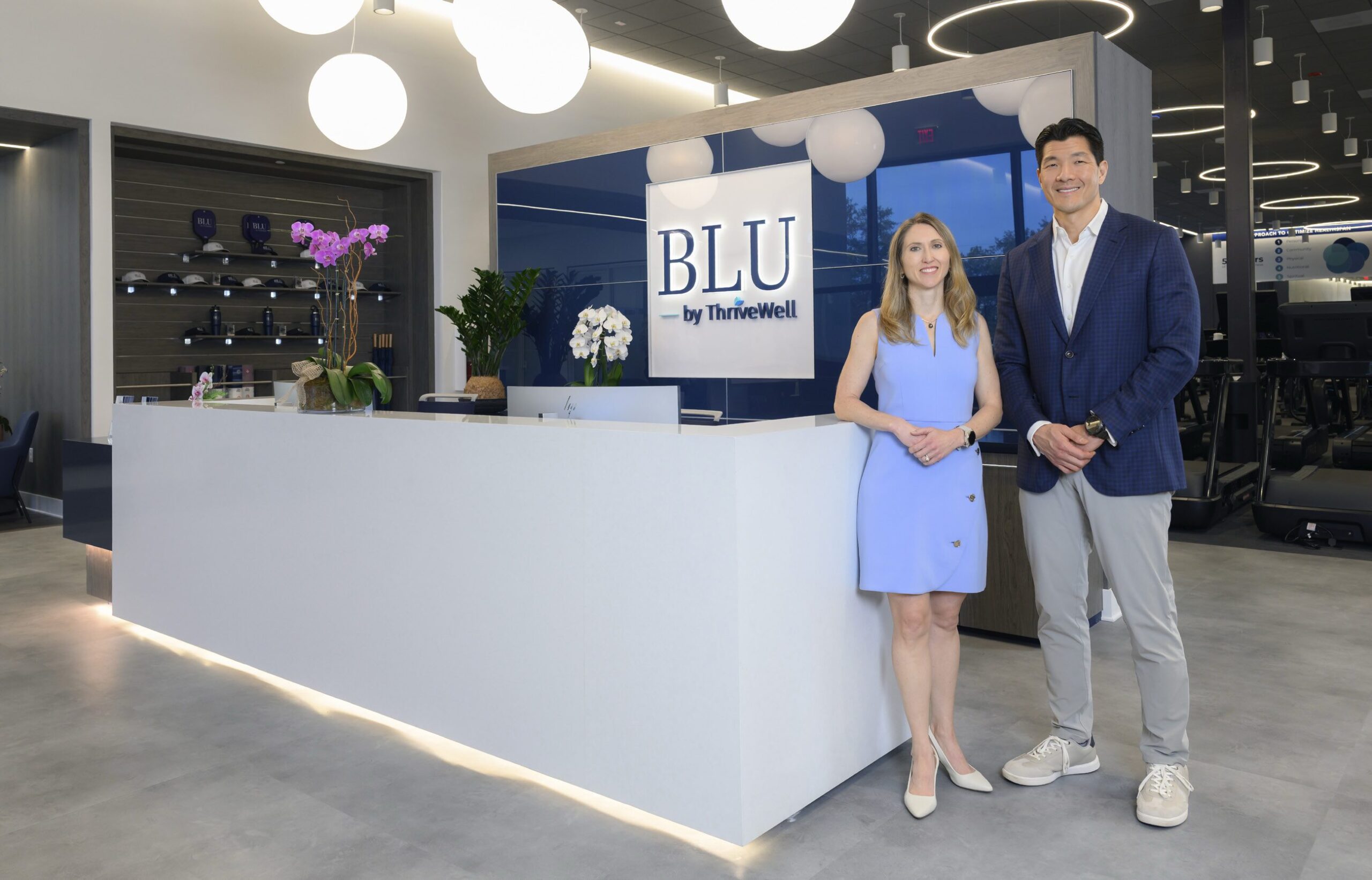 A woman and a man, leaders in Health Care, stand smiling in front of a modern white reception desk with a "BLU by ThriveWell" sign. The room is well-lit, featuring round ceiling lights and contemporary interior design. Lifestyle