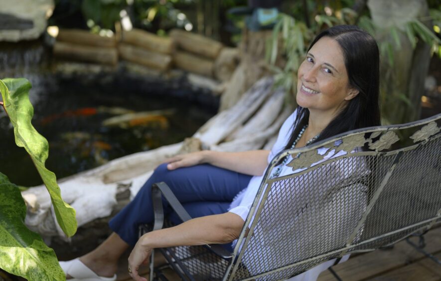 A woman with long dark hair sits on a metal chair in a garden, smiling at the camera. Surrounded by lush greenery and a small pond with koi fish, she radiates confidence—an inspiring presence among health care leaders. Lifestyle