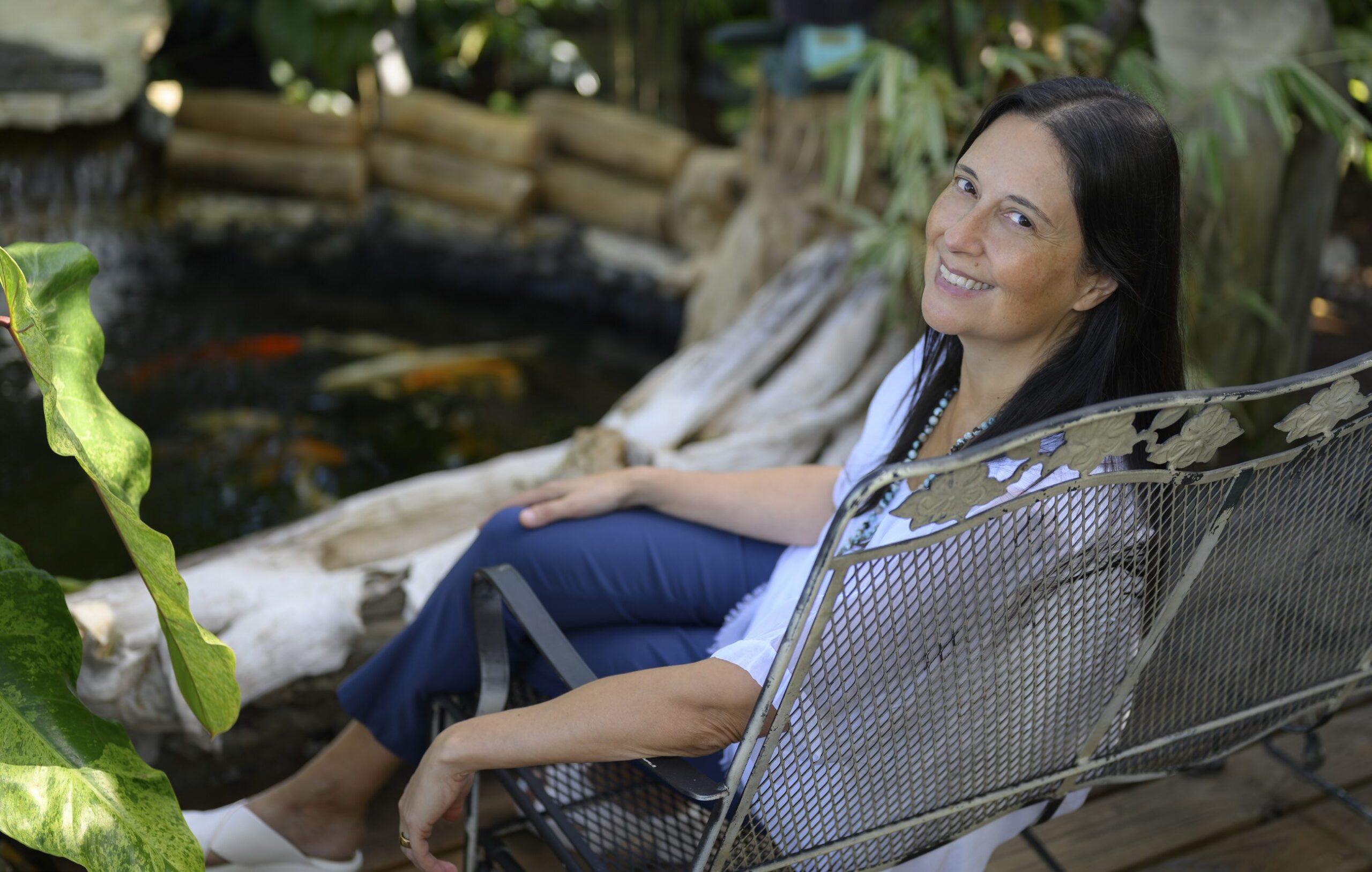 A woman with long dark hair sits on a metal chair in a garden, smiling at the camera. Surrounded by lush greenery and a small pond with koi fish, she radiates confidence—an inspiring presence among health care leaders. Lifestyle