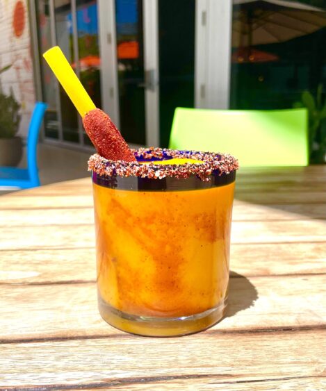 A glass of bright orange drink with chili powder around the rim, a tamarind candy straw, and spicy chili seasoning sits on a wooden table outdoors in sunny Fort Lauderdale weather—a perfect Valentine’s treat. Lifestyle