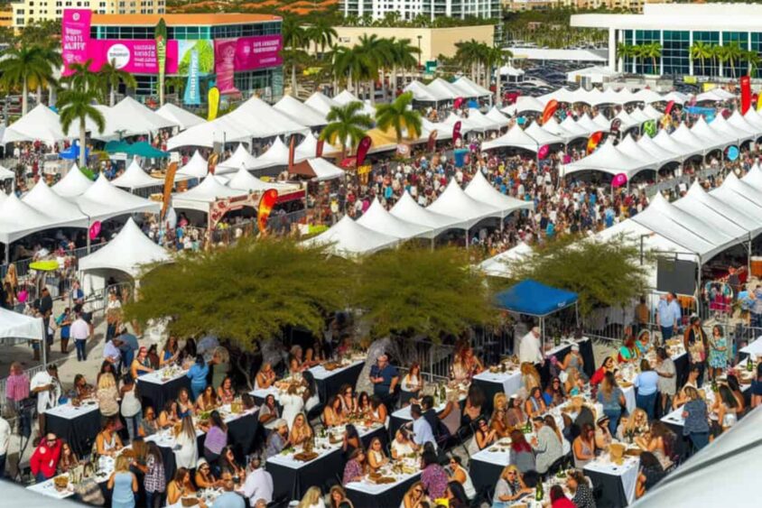 A large outdoor festival in Fort Lauderdale with rows of white tents, crowds walking, and many seated at tables eating and socializing under sunny skies with palm trees—a vibrant celebration of local culture during the week. Lifestyle
