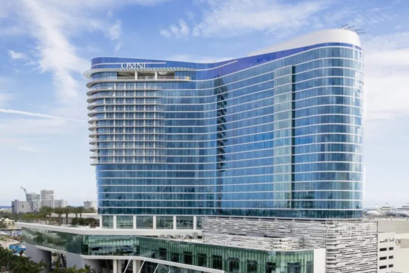 A modern, high-rise glass hotel with curved architecture and multiple balconies, labeled "Omni," stands as a waterfront power play against a blue sky with scattered clouds. Palm trees and city buildings add to the vibrant backdrop. Lifestyle