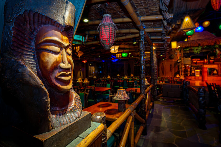 A carved wooden mask with an ornate headdress is displayed in the foreground of a dimly lit, tropical-themed restaurant in Fort Lauderdale, featuring bamboo furniture and colorful hanging lanterns. Lifestyle