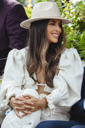 A woman with long wavy hair, wearing a beige wide-brim hat and a white outfit with puffy sleeves, sits outdoors at a relaxed pace, smiling with her hands folded in her lap. Green plants are in the background. Lifestyle
