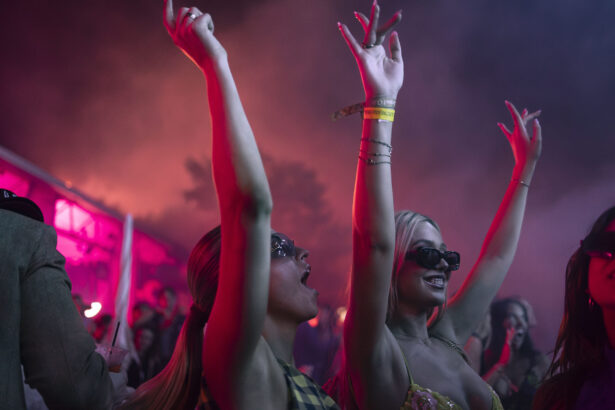 Two women wearing sunglasses cheer and raise their arms while dancing at a lively night event, where Pegasus sets the pace, illuminated by pink and purple lights, with a crowd and smoky atmosphere in the background. Lifestyle