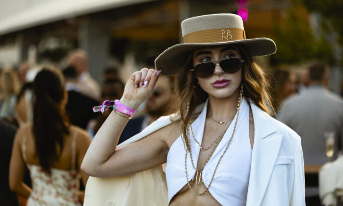 A stylish woman in a white outfit, sunglasses, and a wide-brimmed hat poses confidently at an outdoor event. Channeling Pegasus-like grace, she adjusts her hat and layered necklaces as the lively pace of the crowd blurs in the background. Lifestyle