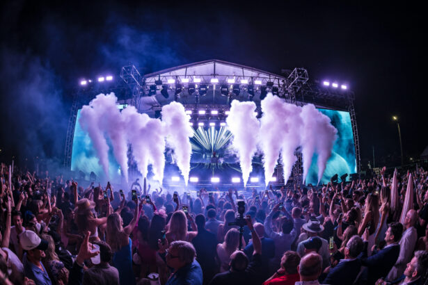 A large crowd enjoys a nighttime concert with bright stage lights, smoke effects, and vibrant visuals on a big screen behind the performers. The audience, celebrating the decade’s biggest hits, holds up phones and cheers near the outdoor stage. Lifestyle