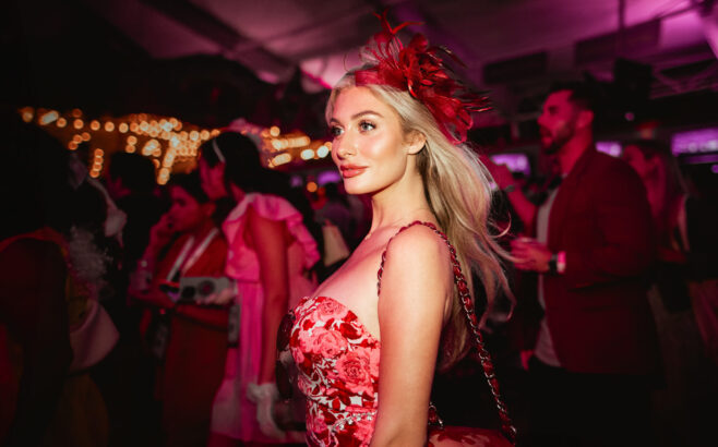 A woman in a red floral dress with a matching red feathered headpiece stands in a dimly lit, festive crowd, illuminated by warm lights and pink hues, as the pace of the celebration recalls the glamour of another decade. Lifestyle