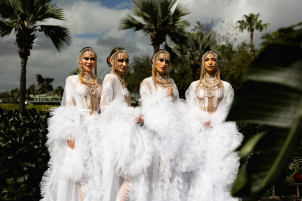 Four women stand outdoors in elaborate white gowns with feathered skirts and gold chain headdresses, evoking a Pegasus-like elegance amid lush greenery and palm trees under a partly cloudy sky. Lifestyle