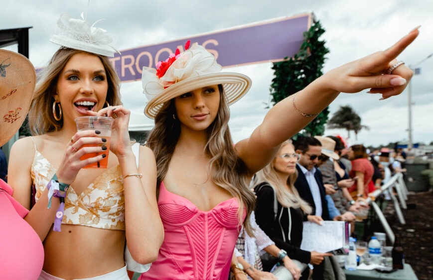 Two women in stylish hats and dresses stand at an outdoor Pegasus event. One drinks a pink beverage and smiles, while the other, in a bright pink top, points excitedly toward something in the distance. People watch behind them. Lifestyle