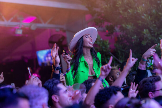 A woman in a bright green outfit and white hat smiles while sitting on someone’s shoulders in a lively crowd at a colorful, outdoor nighttime event. The pace of the celebration is electric as many people cheer and hold up their phones. Lifestyle