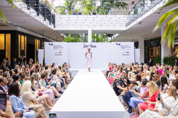 A fashion model walks down a white runway at an indoor fashion show, where Fashion Takes the Lead, surrounded by a large seated audience and "Neiman Marcus" and sponsor logos displayed in the background. Lifestyle