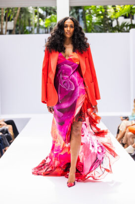 A woman leads the runway in a flowing pink and red floral dress, paired with a bright red blazer and matching heels. She showcases the latest fashion trends with confidence, her long curly hair cascading as greenery frames the white background. Lifestyle