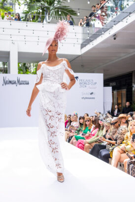 A model in a white lace dress and elaborate pink hat walks a runway at an outdoor fashion show, showcasing the latest fashion trends to a seated audience and onlookers watching from a balcony above. Lifestyle