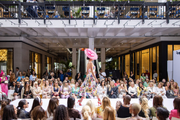 A model wearing a floral dress and large pink hat walks down a runway at an outdoor fashion show, showcasing the latest fashion trends to a large seated audience and onlookers watching from an upper balcony. Lifestyle