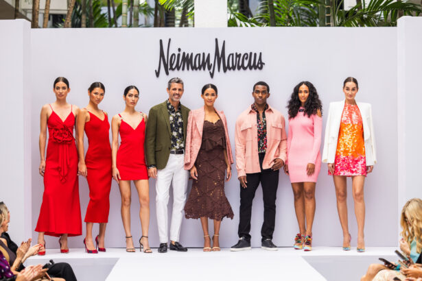 A diverse group of models and two individuals in the center pose on a white runway before a Neiman Marcus sign, showcasing vibrant spring and summer outfits that highlight the latest fashion trends in red, pink, brown, and white tones. Lifestyle