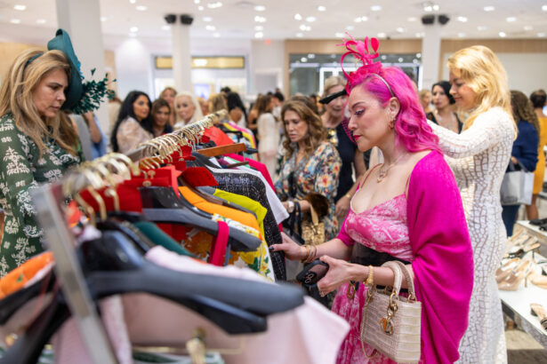 Women browse colorful clothing on racks at a busy, well-lit boutique. One woman with bright pink hair and a matching fascinator stands out, showcasing fashion leadership in a pink and floral dress while holding a small purse. Lifestyle