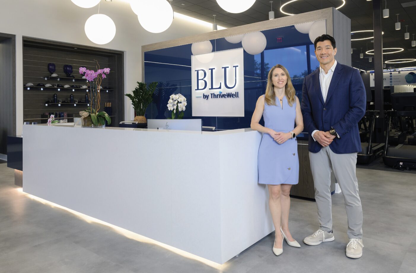A woman and a man stand smiling at the reception desk of a modern wellness center with the sign "BLU by ThrIVeWell" on the wall behind them, showcasing a shift from sick care to a strategy focused on proactive well-being in this bright, stylish space. Lifestyle