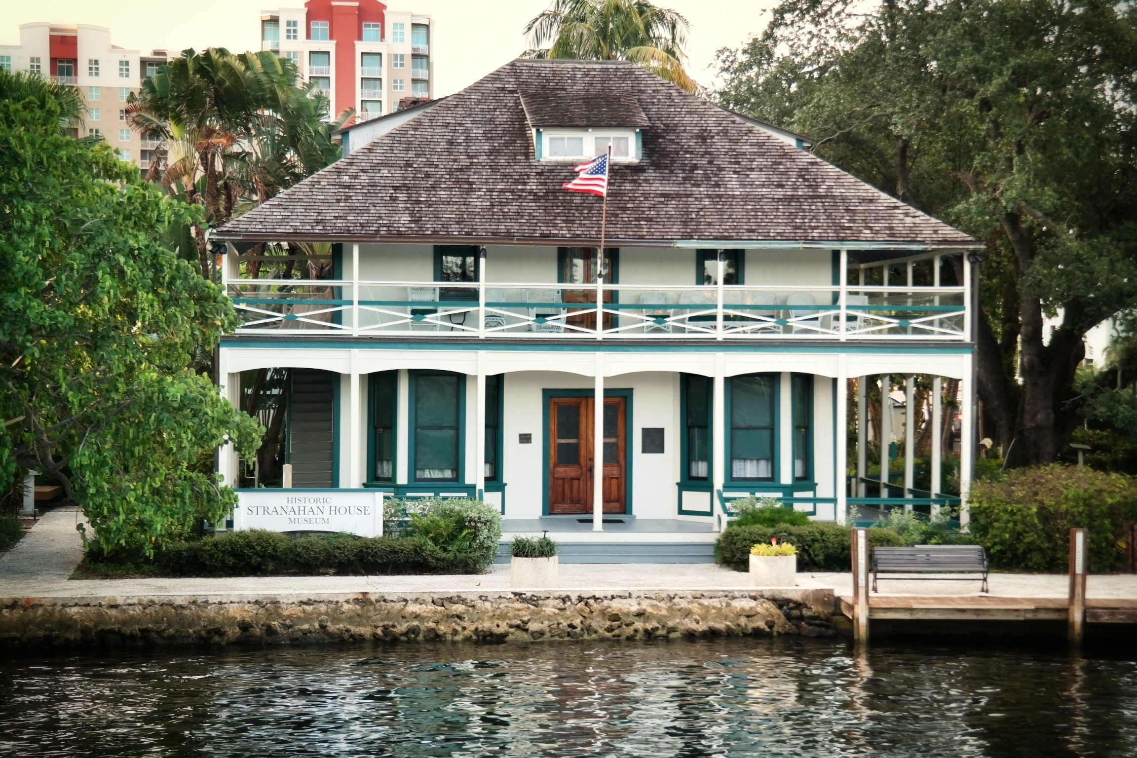 A historic two-story house with a wooden porch and American flag sits by the river, surrounded by trees. A sign in front reads “Historic Stranahan House Museum,” with modern buildings visible in the background. Lifestyle